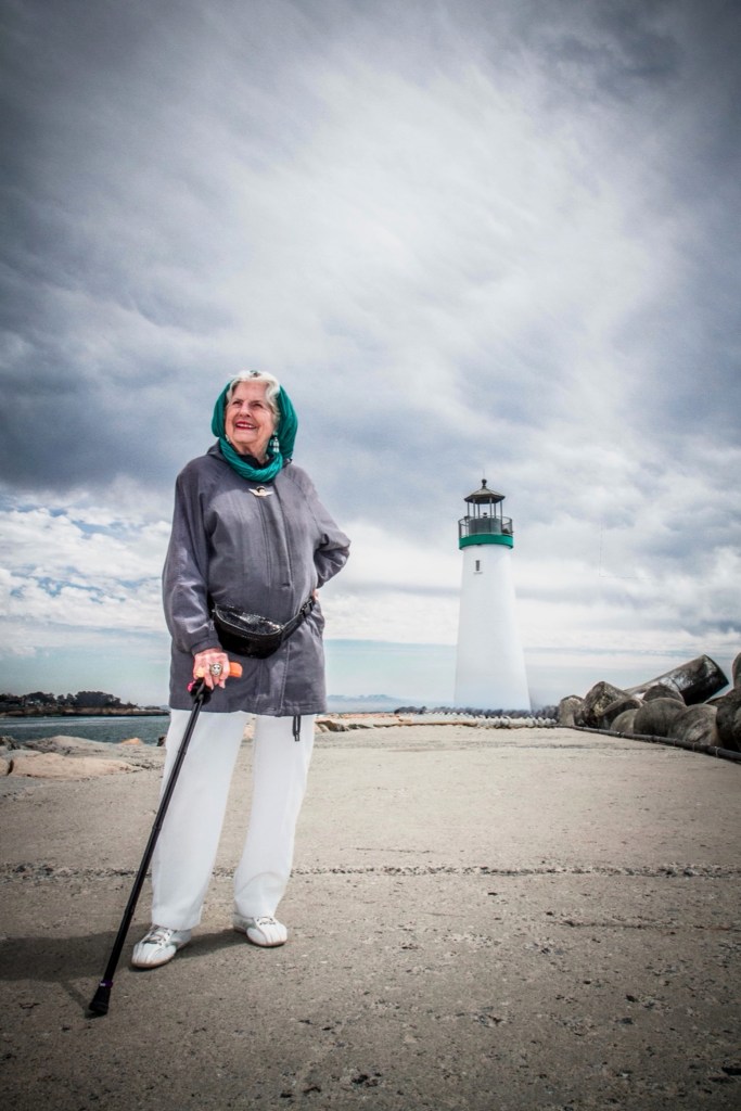 image of Betty Ann, elderly white woman, looking regal in a blue jacket and turquoise head scarf, hand on hip, gazing into the distance, smiling radiantly. one hand holds her cane, behind her, the Santa Cruz lighthouse is visible. The sky is cloudy, adding a sense of drama.